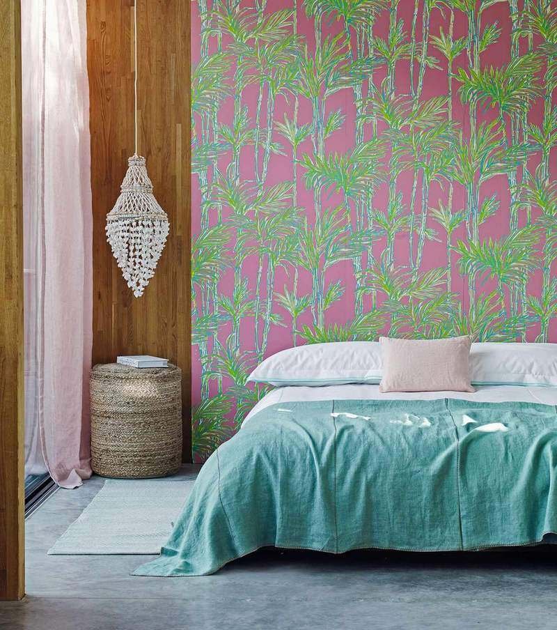 A colorful bedroom with a pink and green patterned statement wall, wood panelling, a macrame hanging, rattan stool, and a low bed with blue bedding.
