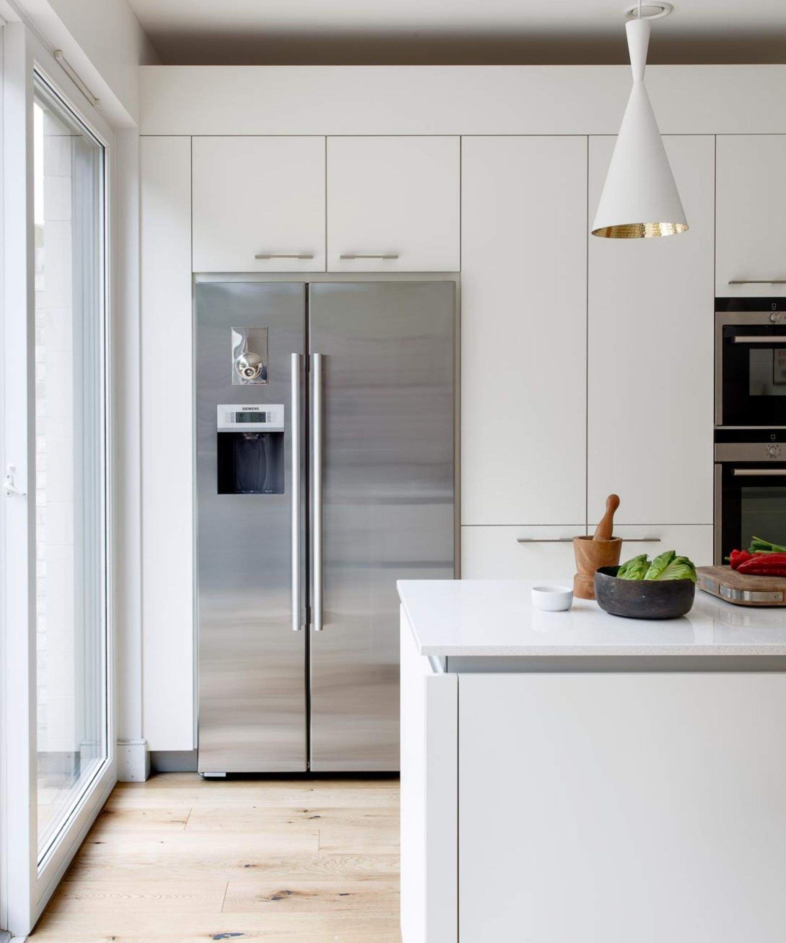 Stainless steel American style fridge freezer in a wall of floor-to-ceiling kitchen cabinets. The aesthetic of the kitchen is white, clean and minimalist with wooden floors and large kitchen island with spot lighting