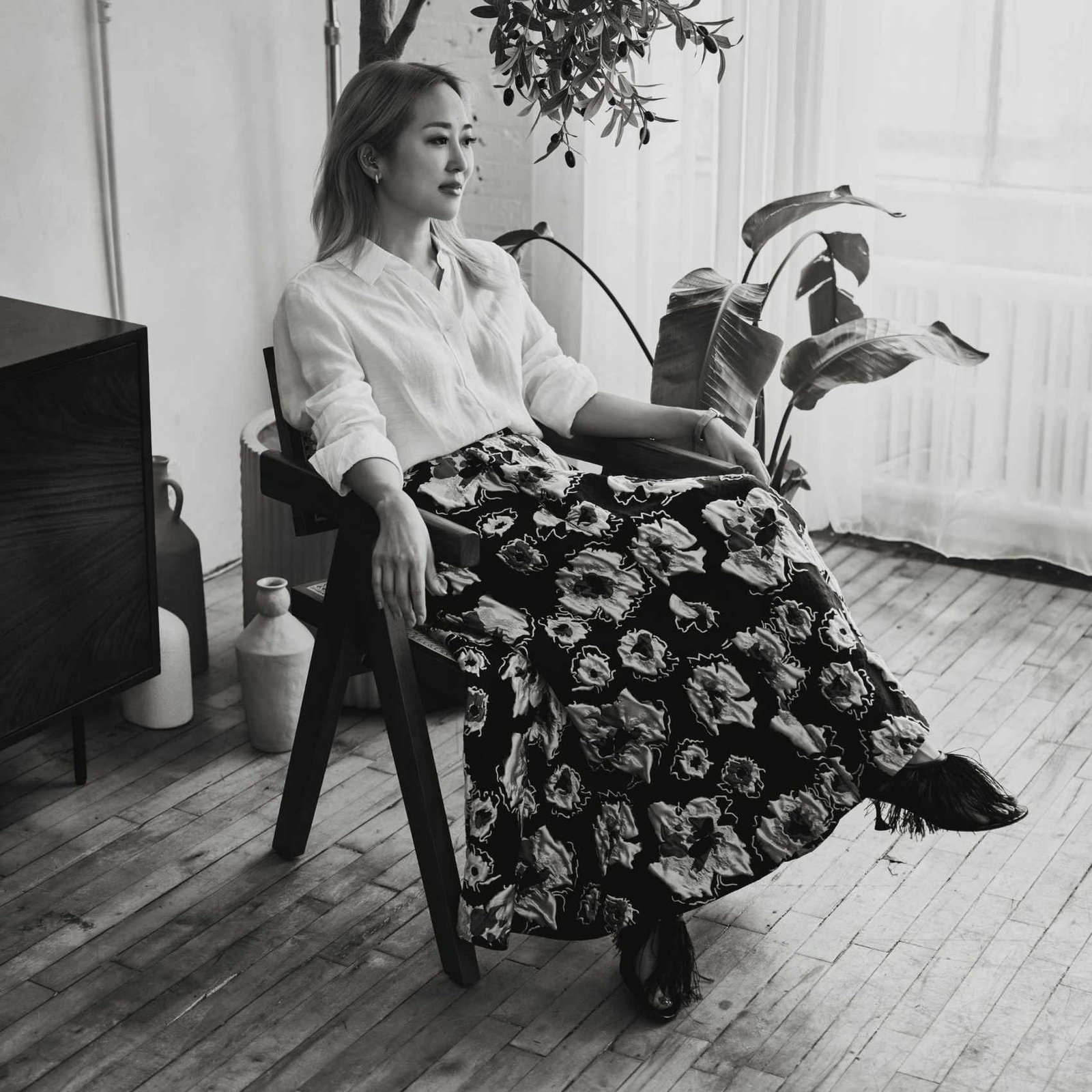 A black and white image of Maiko Shimazaki wearing a white skirt and black and white patterned skirt, sitting in a chair.