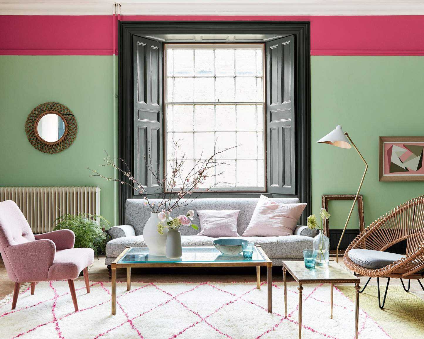 Bright, colorful living room with green painted walls and pink painted trim, large window with wooden shutters, light wooden flooring, cream and pink rug, gray sofa, pink armchair, wicker style armchair, wooden and glass coffee table, artwork and mirrors on wall
