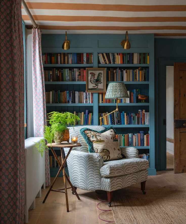 A corner of a room with a floor to ceiling bookshelf painted in a dark blue colour. The bookshelf is full of books and on the centre panel hangs a small painting of a cockerel. A patterned armchair is in front of it with a large cushion with a blue tassel trim. A small wooden table sits beside it with a plant pot and a candle on top.