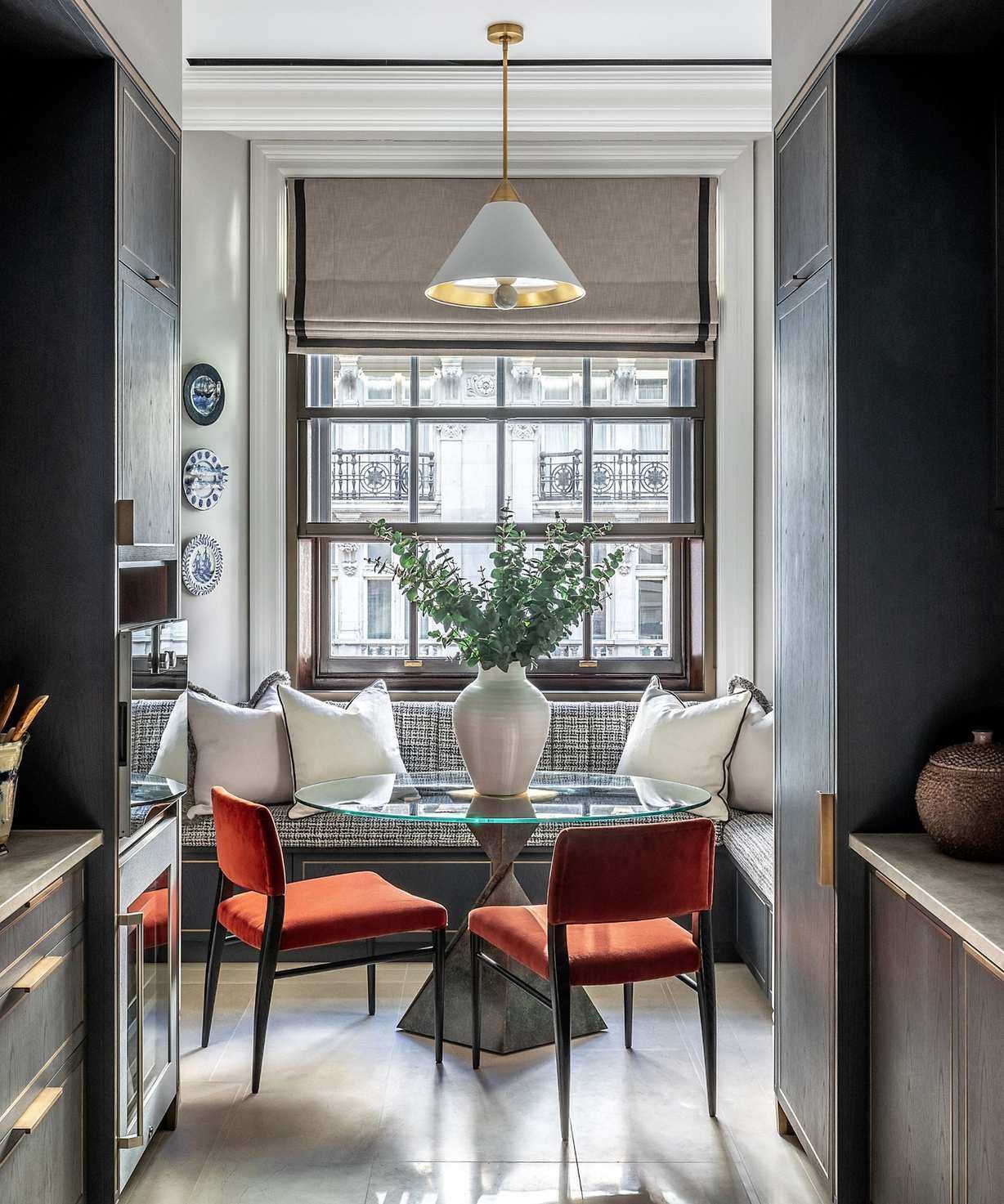 A small round glass kitchen dining table in front of a banquette bench, with two red velvet chairs tucked beneath. In a large window at the end of a narrow kitchen.