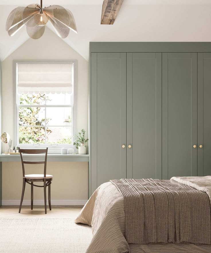 Cream bedroom with carpet, sage green built-in closet and matching vanity table with mid century chair. There is a petal statement ceiling light and bright window showing trees beyond
